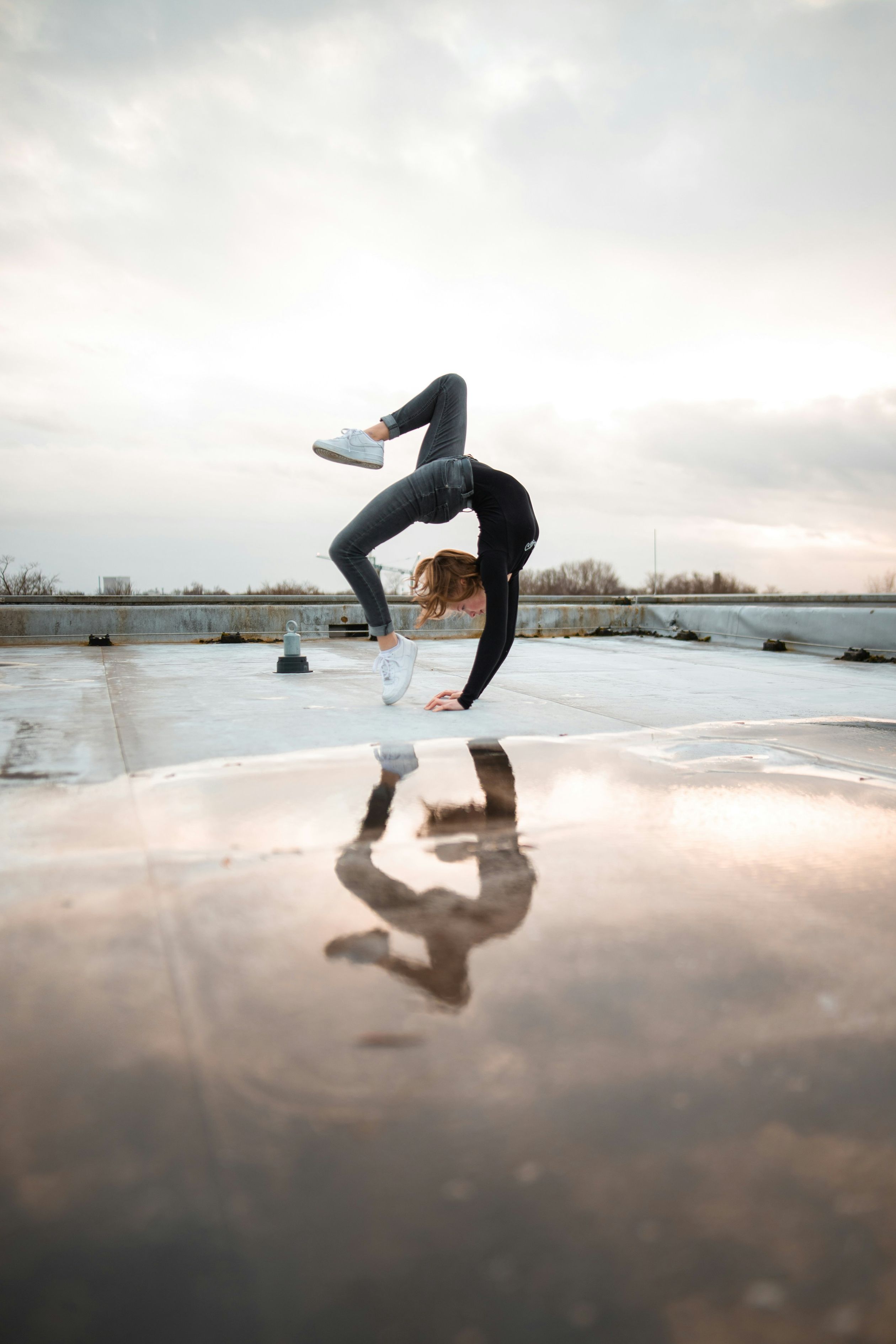 Person performing a scorpion handstand on a wet rooftop, with their reflection visible in a puddle.