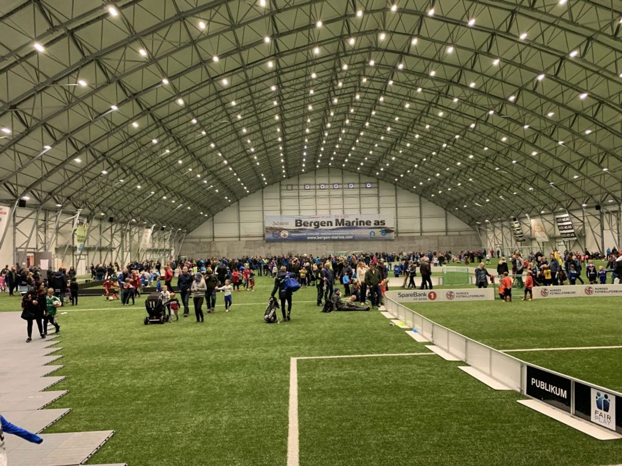 A large indoor sports arena with an artificial turf field, crowded with adults and children under a brightly lit arched ceiling.