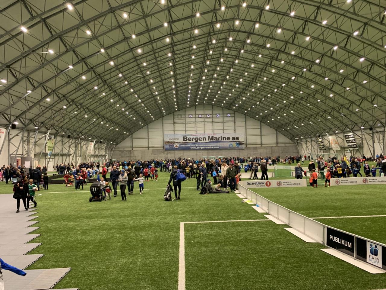 A large indoor sports hall with artificial turf, filled with many families and children under a high, arched ceiling.