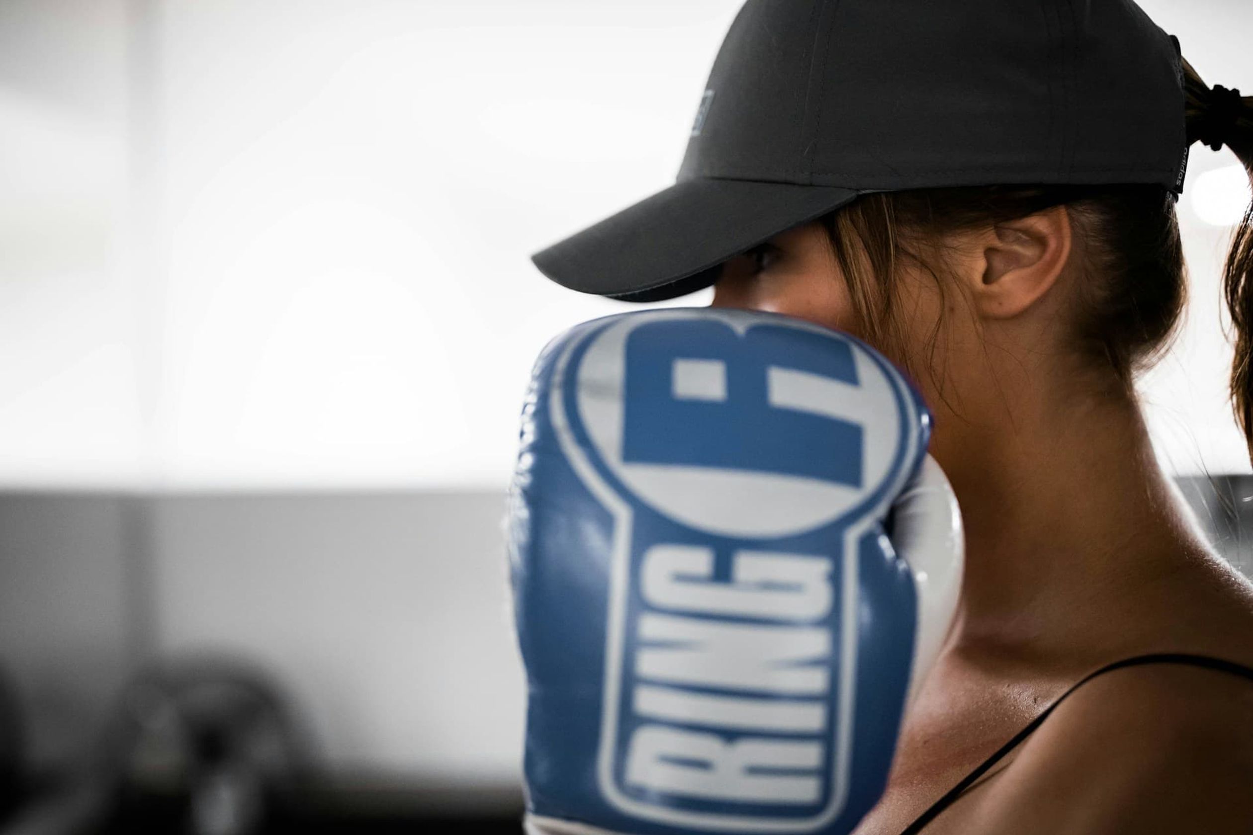 Woman in grey cap and blue 'RING' boxing glove covering her face.