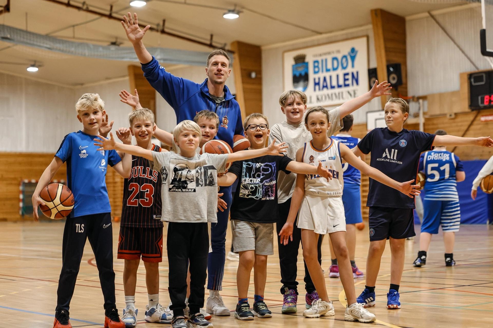 A basketball coach and a group of happy children pose with outstretched arms on a court.