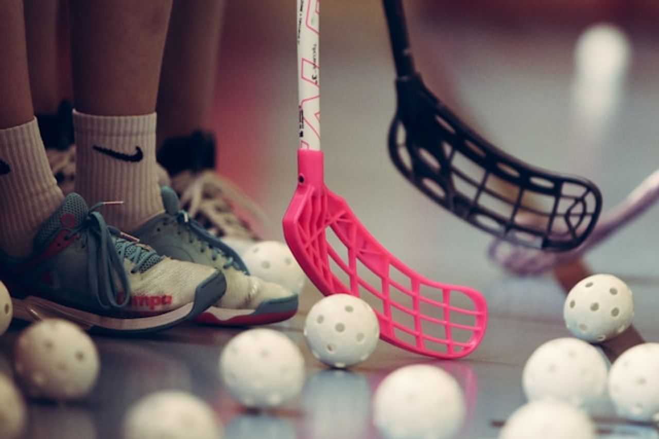 Close-up of floorball players' feet, pink and black sticks, and white balls on a court.