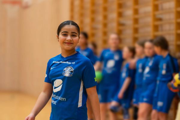 A young girl in a blue sports jersey smiles at the camera in an indoor gym, with blurred children in the background.