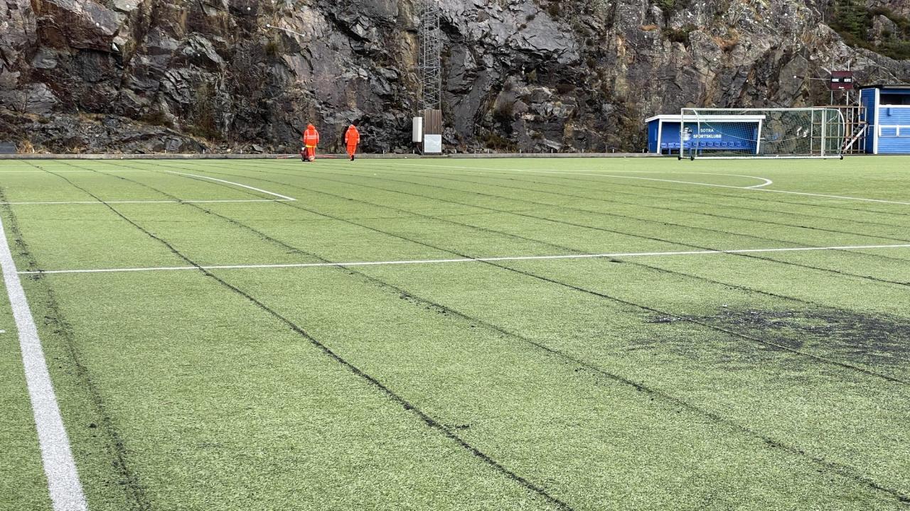 An artificial turf sports field with two workers in orange vests, a rocky hill, and a soccer goal in the background.