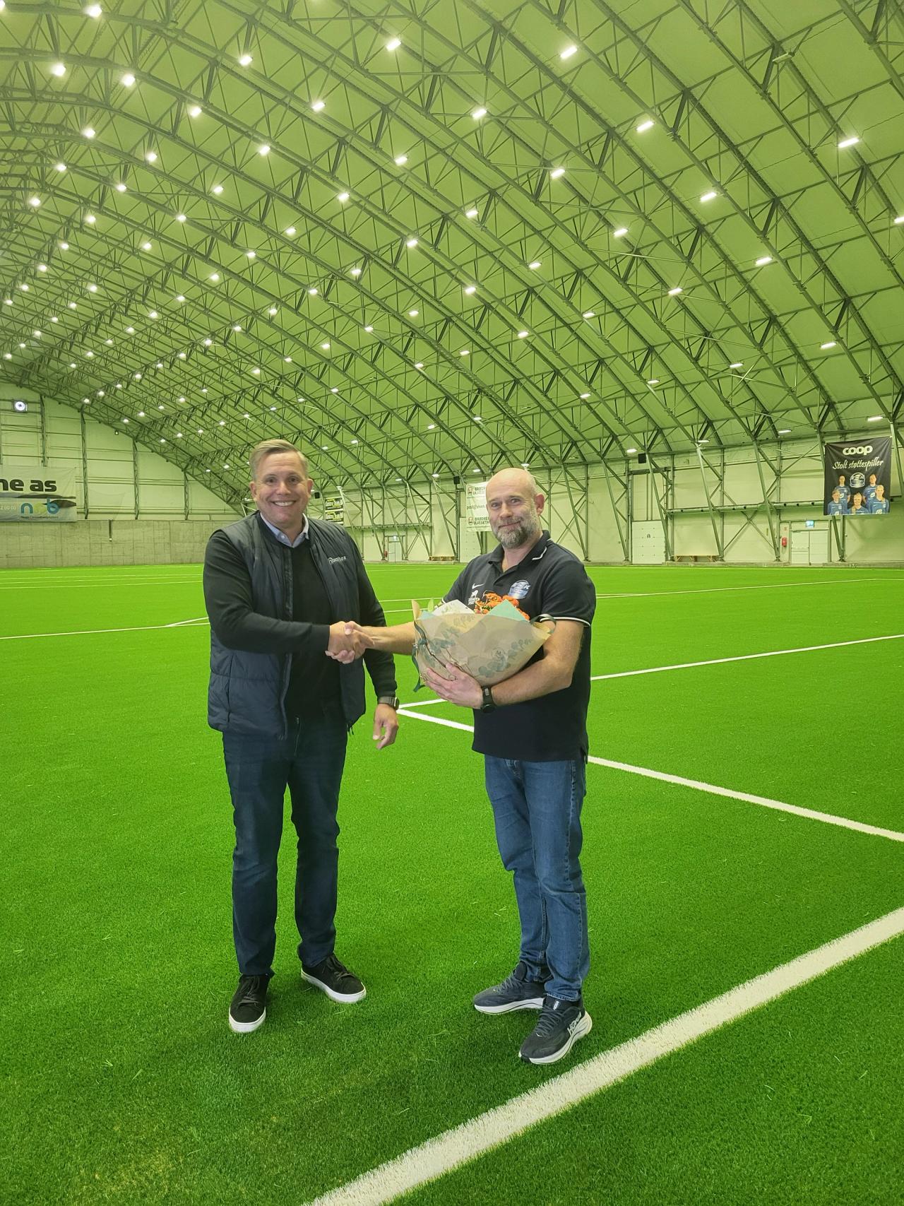 Two men shake hands on an indoor artificial turf field, one holding flowers.