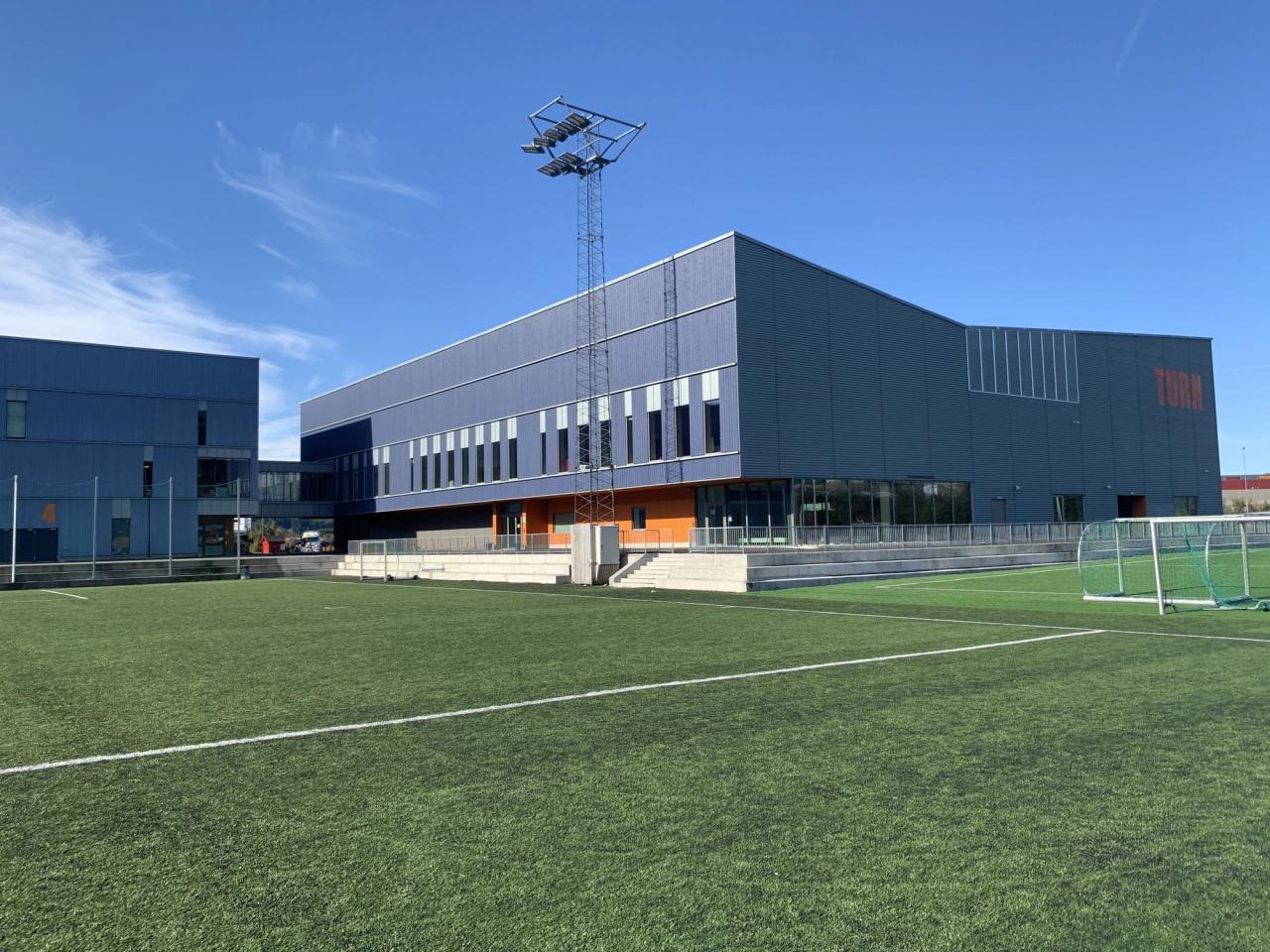 Modern blue building next to an artificial turf sports field with a soccer goal and a light pole under a clear sky.
