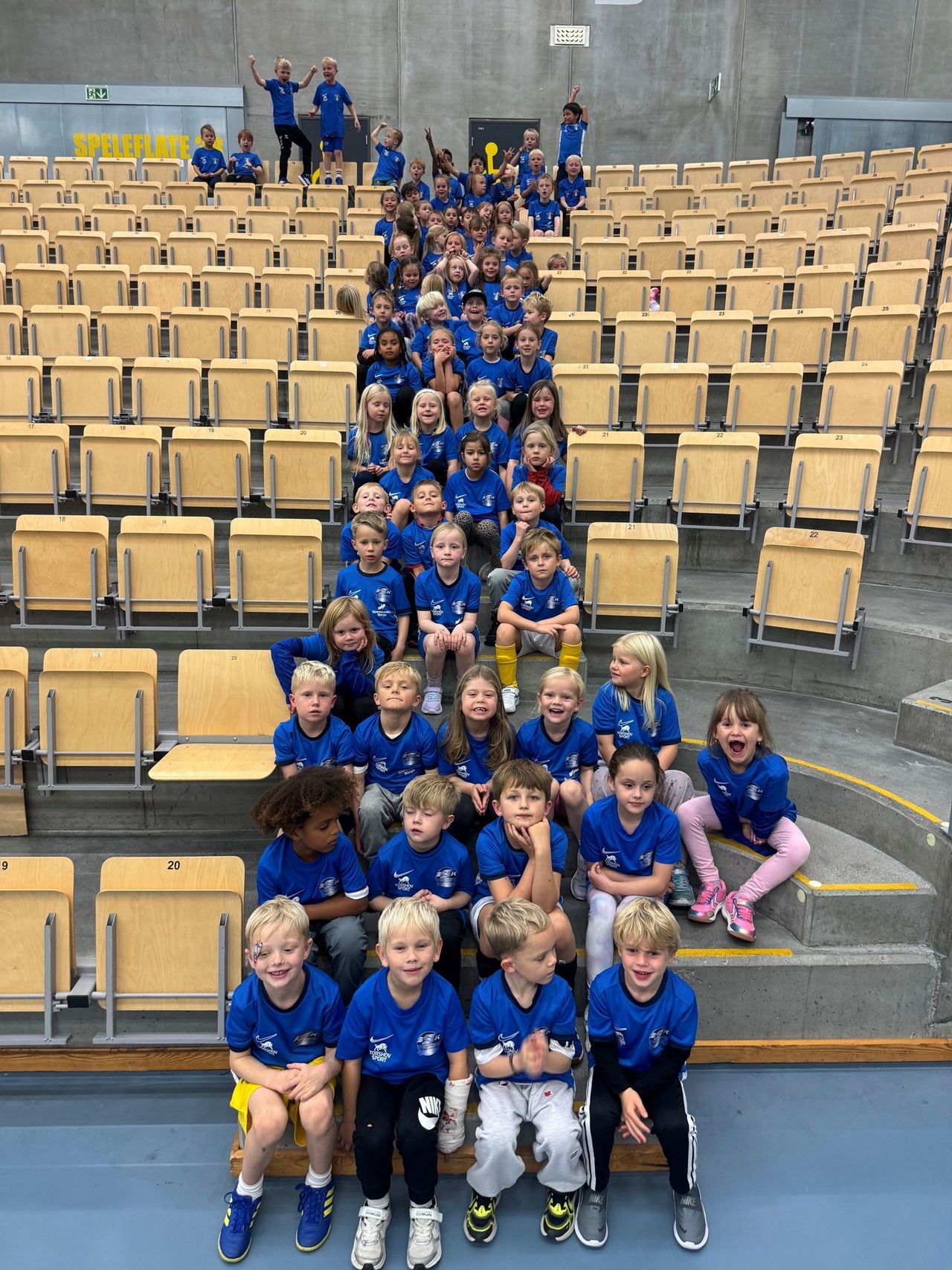 A large group of children and some adults in blue uniforms pose on tiered stadium seating.
