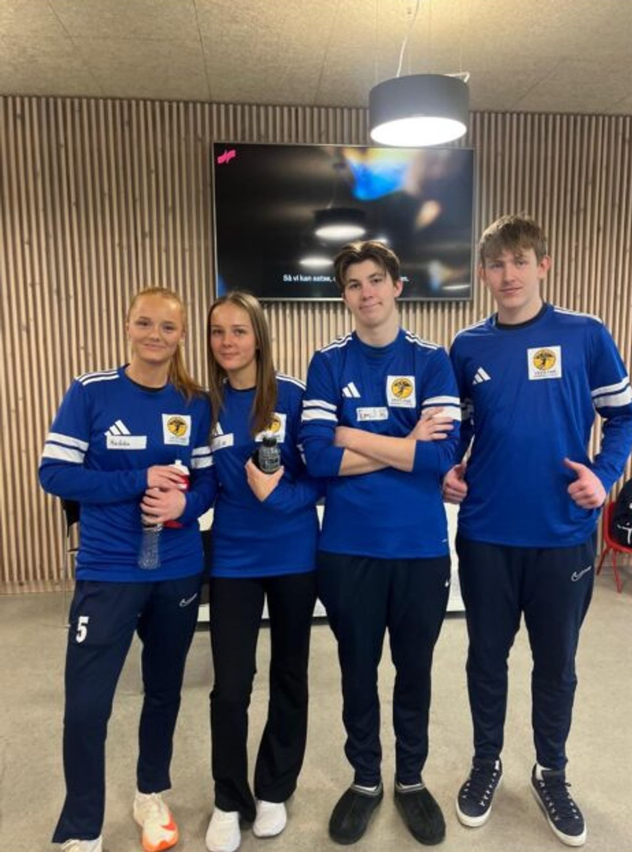 Four young people in matching blue sportswear with yellow logos stand in front of a wooden wall and a TV screen.