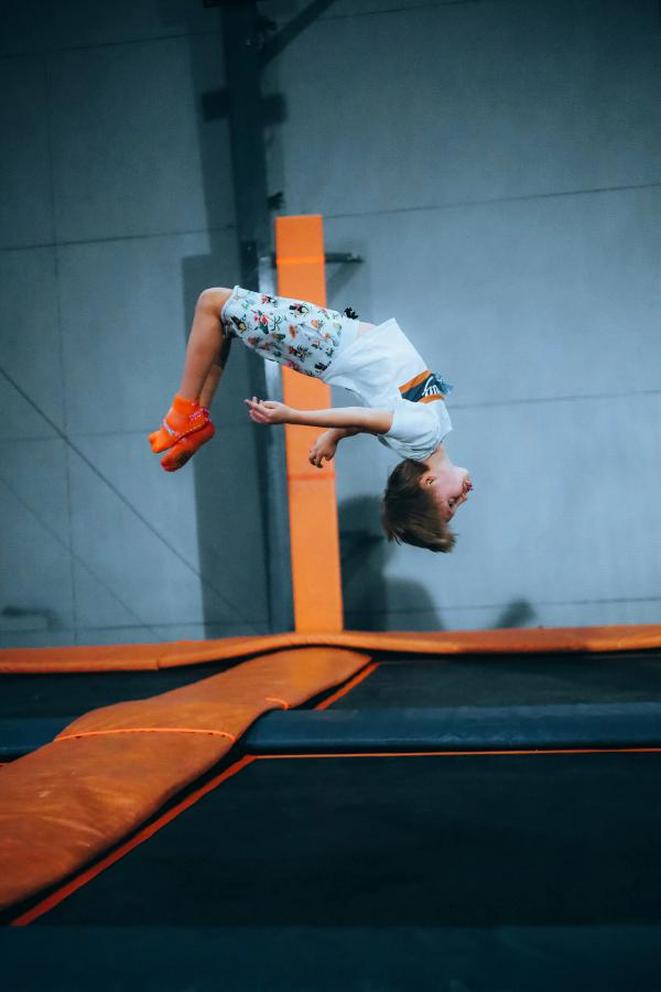 A young boy in patterned shorts and orange socks flips upside down in a trampoline park.