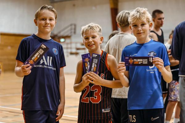 Three boys smile, holding Fyllingen Lions tickets on a basketball court.