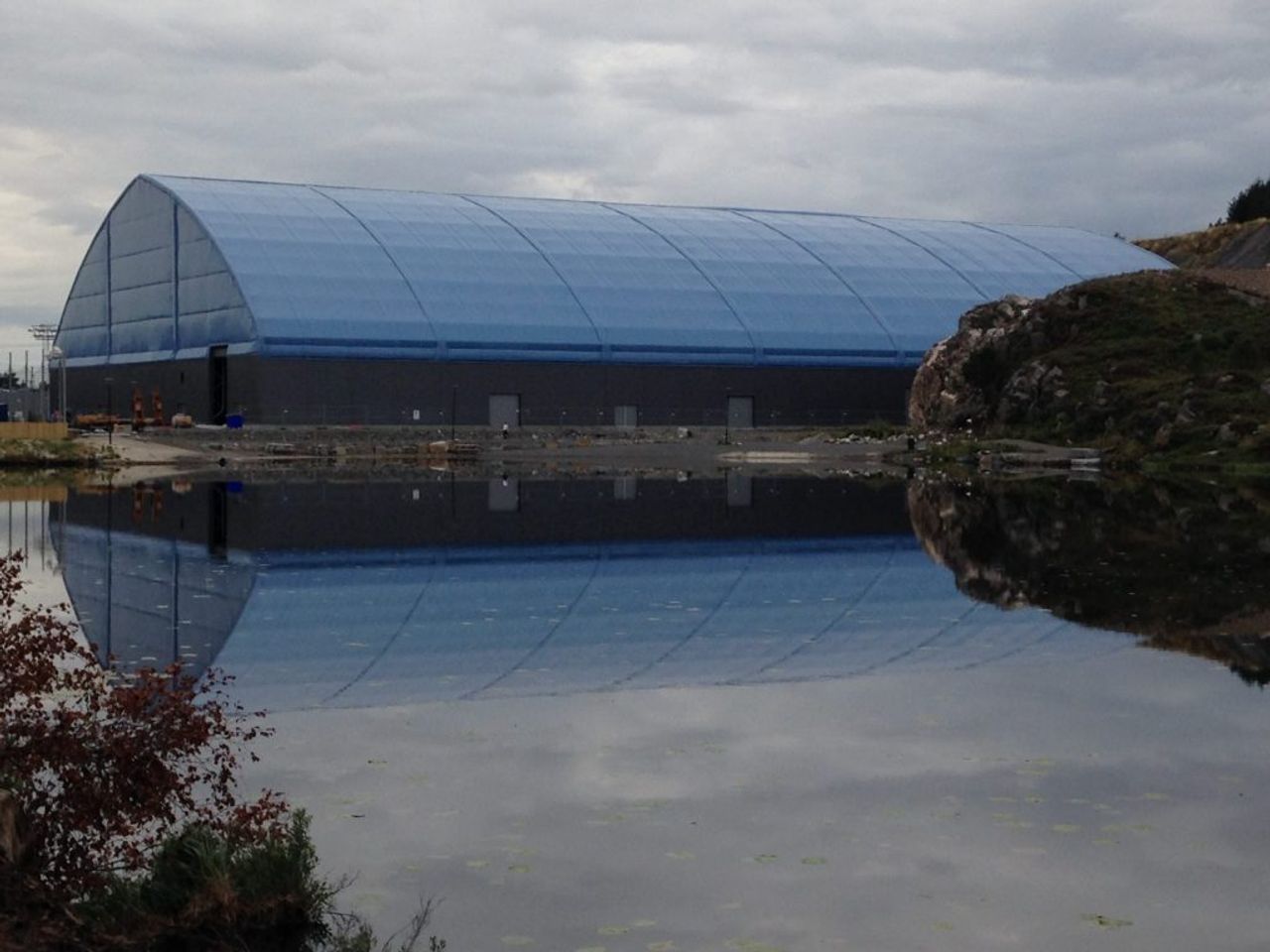 A large, long building with a blue arched roof and dark grey walls, reflected in calm water with a rocky hillside on the right.