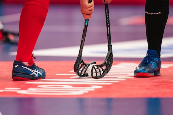 Two floorball players face off with sticks over a ball on a red court.