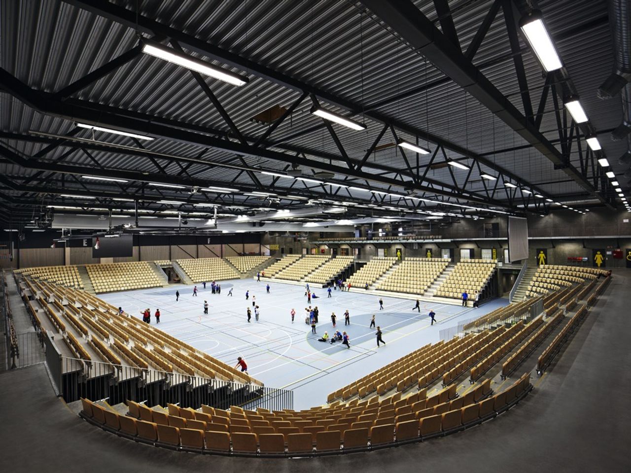 A modern indoor sports hall with a blue court where many children are playing, surrounded by tiered seating.