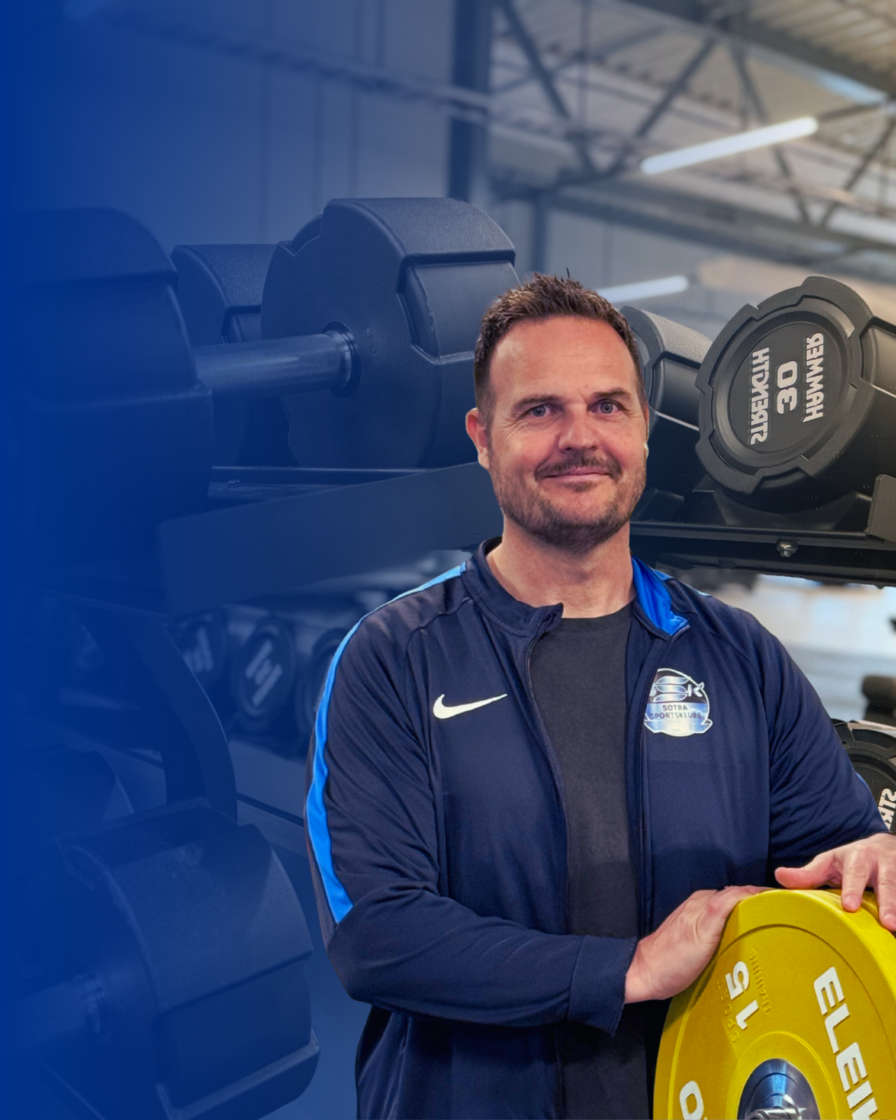 Smiling man in a navy tracksuit holding a yellow weight plate in a gym.