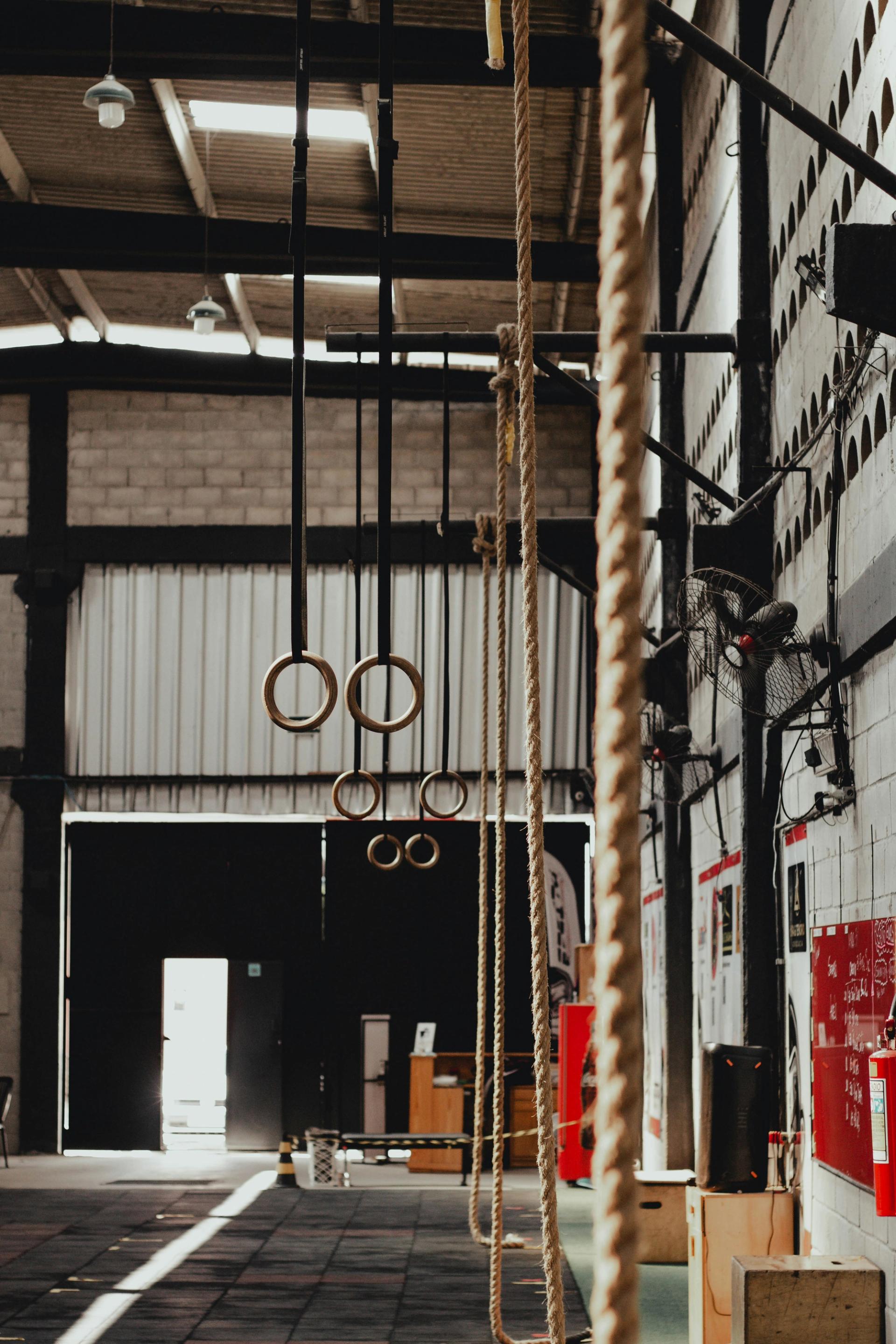 Industrial gym with hanging climbing ropes, gymnastic rings, and a bright doorway in the background.