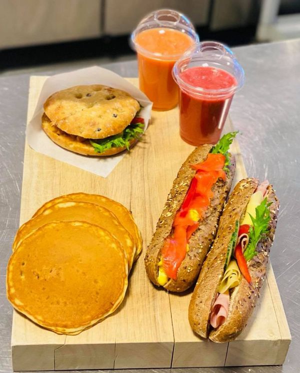 A wooden board displays a sandwich, two sub rolls, three pancakes, and two fruit smoothies.