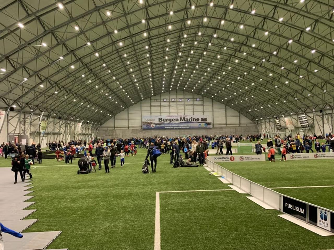 An indoor sports hall with artificial turf, a domed ceiling, and a large crowd of adults and children.