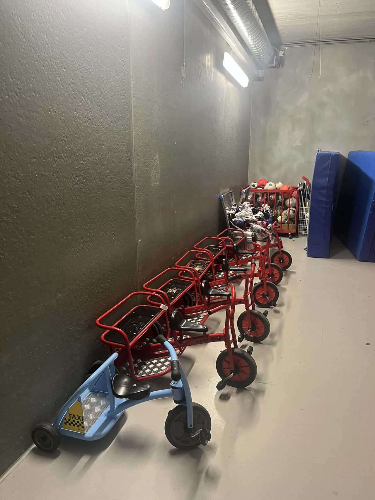 A line of children's tricycles, including a blue taxi, against a wall in a room with play equipment.
