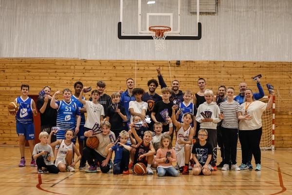A group of children, young adults, and adults smiling on a basketball court, many holding prizes.
