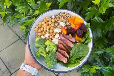 A hand holds a colourful bowl with sliced lamb, broccoli, chickpeas, quinoa, roasted vegetables, spinach, and feta.