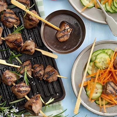 Grilled meatmb skewers with rosemary, one dipping into sauce, and plates of cucumber and carrot salad. Grilled meatmb skewers with rosemary, one dipping into sauce, and plates of cucumber and carrot salad.