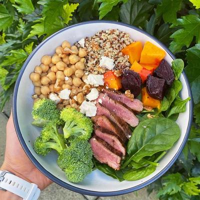 A colorful bowl with sliced lamb, broccoli, chickpeas, quinoa, roasted squash and beets, spinach, feta, and walnuts.