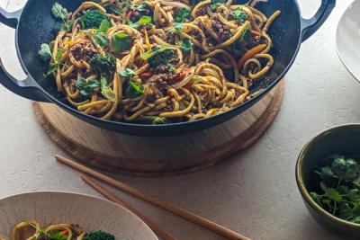 A black wok filled with stir-fried noodles, ground lamb, broccoli, vegetables, and fresh herbs.