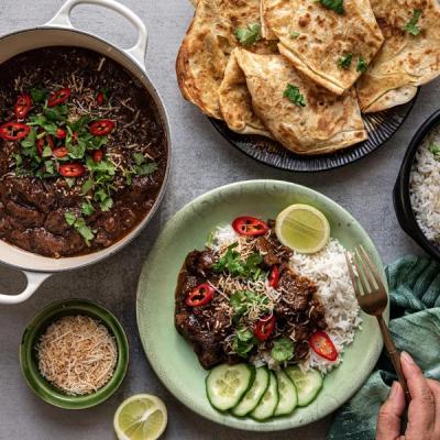 A dark, rich curry, garnished with chili, cilantro, and toasted coconut, served in a pot and on a plate with rice, cucumber, and lime, next to flatbreads.