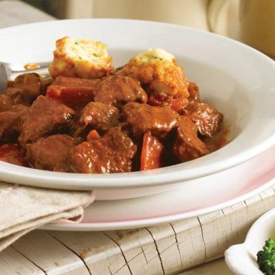 A white bowl of hearty beef stew with chunks of meat, vegetables, and two savory dumplings on top.