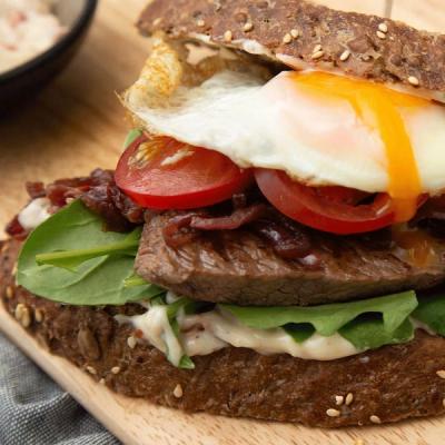 A steak sandwich on whole-grain bread with a fried egg, runny yolk, tomato, spinach, caramelized onions, and creamy sauce.