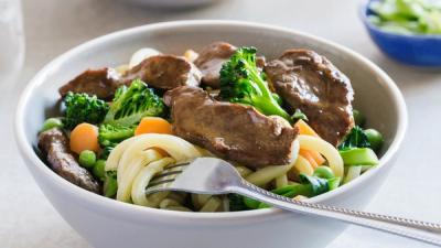 A bowl of udon noodle stir-fry with beef, broccoli, carrots, and peas, with a fork. A bowl of udon noodle stir-fry with beef, broccoli, carrots, and peas, with a fork.
