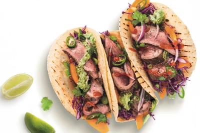 Three lamb tacos filled with purple cabbage, red onion, jalapeños, cilantro, and sauces, next to lime wedges on a white background. Three lamb tacos filled with purple cabbage, red onion, jalapeños, cilantro, and sauces, next to lime wedges on a white background.