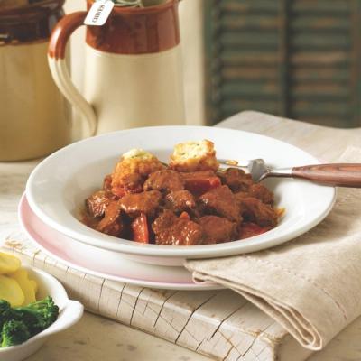 A white bowl of beef stew topped with dumplings, served with a fork on a rustic wooden surface.