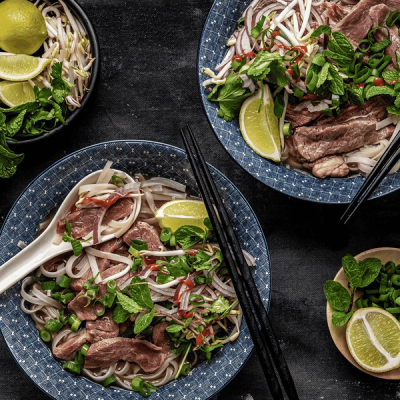 Two bowls of beef pho with noodles, herbs, bean sprouts, and lime slices on a dark background.