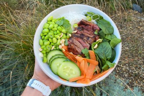 A hand holds a bowl of sliced grilled steak, edamame, cucumber, carrot ribbons, spinach, rice vermicelli, and chopped peanuts. A hand holds a bowl of sliced grilled steak, edamame, cucumber, carrot ribbons, spinach, rice vermicelli, and chopped peanuts.