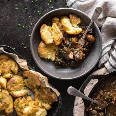 A bowl of beef stew and crispy smashed potatoes, with more potatoes in a pan and a skillet of stew.