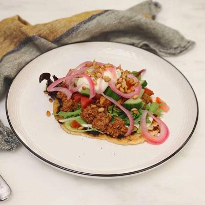 A flatbread topped with spiced meat, mixed salad, pickled red onions, white sauce, and pine nuts on a white plate. A flatbread topped with spiced meat, mixed salad, pickled red onions, white sauce, and pine nuts on a white plate.