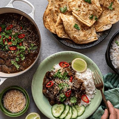 a person is holding a fork over a plate of beef rendang with rice and cucumber on the side on a table . a person is holding a fork over a plate of beef rendang with rice and cucumber on the side on a table .