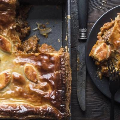 A golden-crusted savory pie in a baking dish, with a slice on a black plate, knife, and fork on a dark wooden table. A golden-crusted savory pie in a baking dish, with a slice on a black plate, knife, and fork on a dark wooden table.