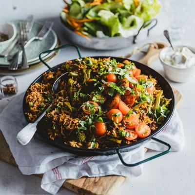 A black paella pan of rice, broccoli, and ground meat, topped with cherry tomatoes and herbs, with a side salad and dip.