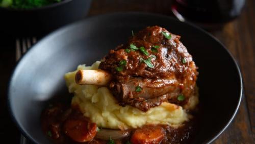 Braised lamb shank on mashed potatoes with gravy and carrots in a dark bowl. Braised lamb shank on mashed potatoes with gravy and carrots in a dark bowl.