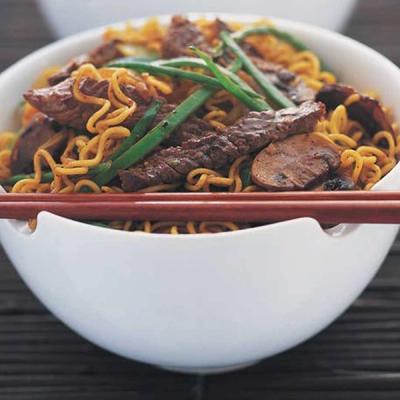 A white bowl of beef and curly noodle stir-fry with green beans, mushrooms, and chopsticks resting on the rim.