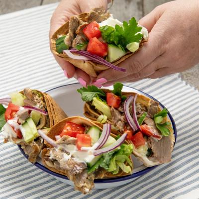 Hands holding a small flatbread wrap filled with shaved meat, fresh vegetables, and sauce, with three more on a plate. Hands holding a small flatbread wrap filled with shaved meat, fresh vegetables, and sauce, with three more on a plate.