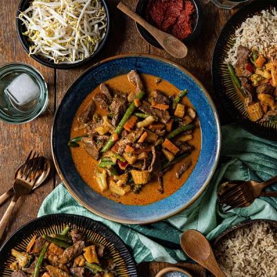 a bowl of beef curry with vegetables and rice on a wooden table . a bowl of beef curry with vegetables and rice on a wooden table .