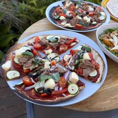 Two rustic flatbread pizzas topped with leftover roast lamb, tomatoes, zucchini, and mozzarella, served with a pear and walnut salad on an outdoor wooden table.