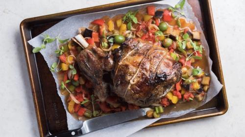 Roasted meat joint with colorful vegetables, green olives, and fresh herbs on a baking sheet.