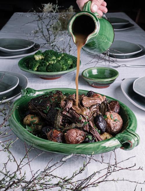 a person is pouring gravy into a green bowl of roast meat and potatoes on a table.