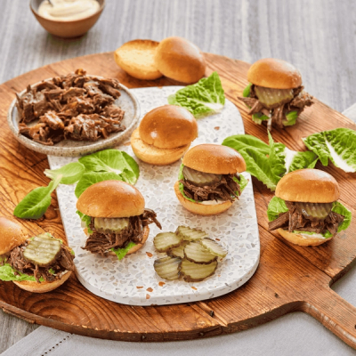 A wooden board holds a white serving tray featuring several mini beef sliders, some fully assembled with lettuce and pickles, alongside additional shredded beef, buns, lettuce, and sliced pickles.