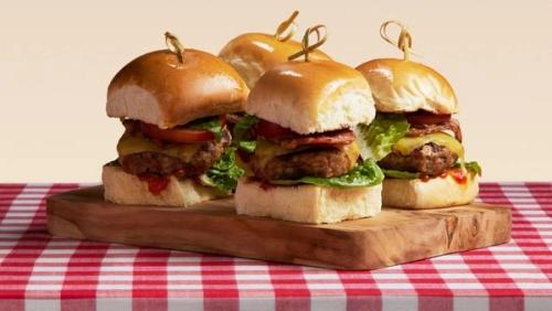 Four gourmet sliders on a wooden board placed on a red and white checkered tablecloth. Four gourmet sliders on a wooden board placed on a red and white checkered tablecloth.