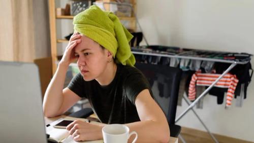 A young woman with a green towel on her head, looking stressed at a laptop on a desk.
